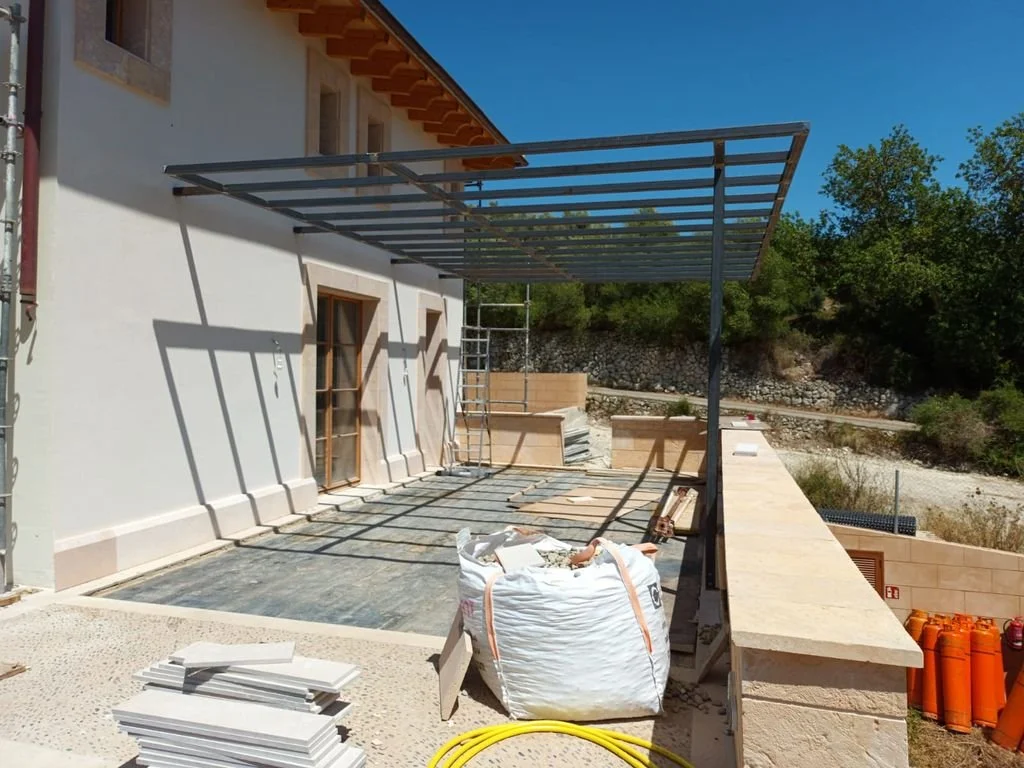 Estructura metálica en construcción en una terraza de una casa, con paredes blancas y ventanas de madera, rodeada de naturaleza y herramientas de construcción.