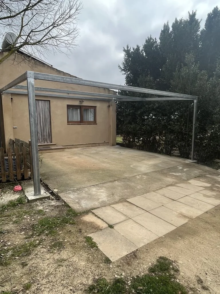 Estructura metálica en un patio cercado con una casa de fondo, cielo nublado, con algunos árboles y pavimento de concreto y piedras.