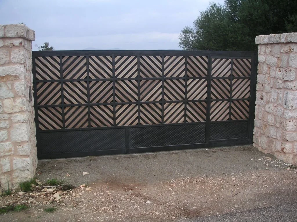 Una puerta de metal decorativa con un patrón de líneas diagonales de color beige y negro, entre dos muros de piedra, de color claro.
