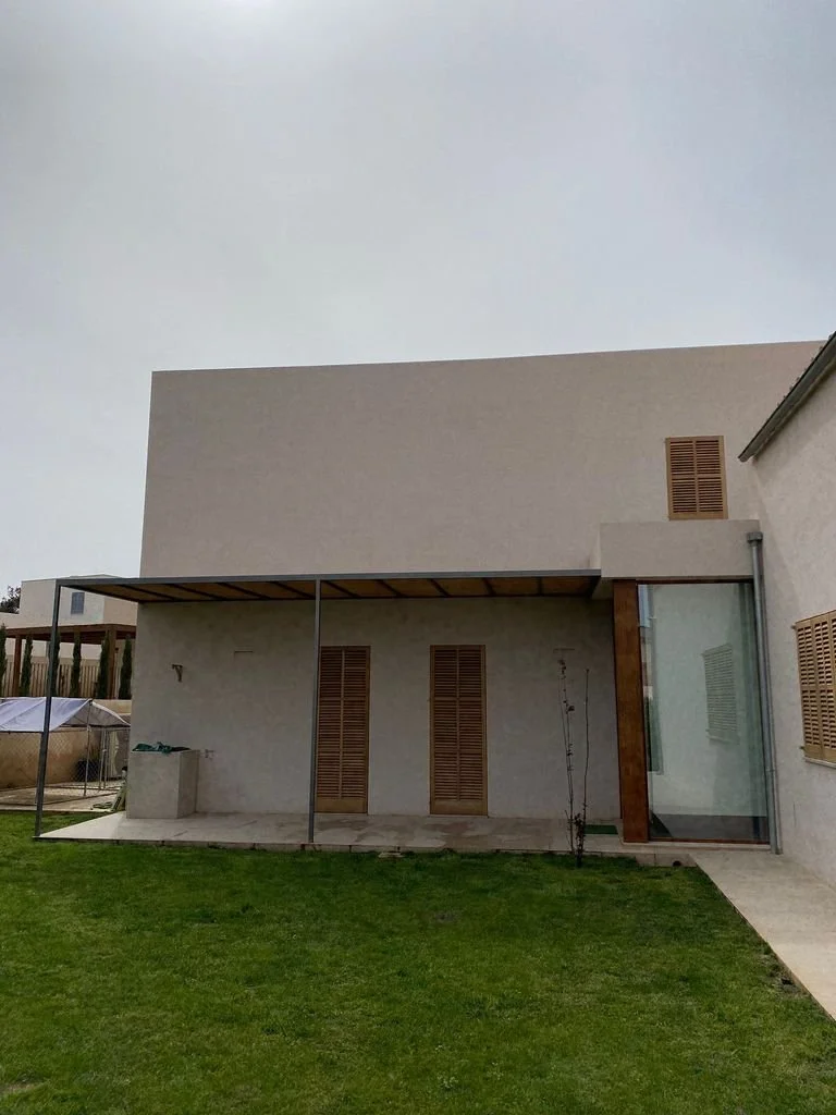 Vista del exterior de una casa moderna con pared blanca, ventanas con persianas de madera y un área de patio con césped y una estructura metálica.