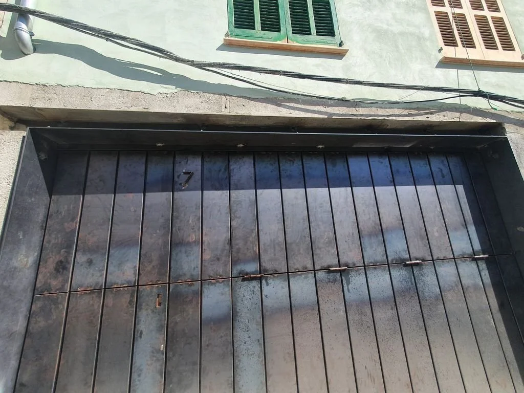 Puerta de garaje de metal oxidado con el número 7, en una calle con cables eléctricos y ventanas con persianas.