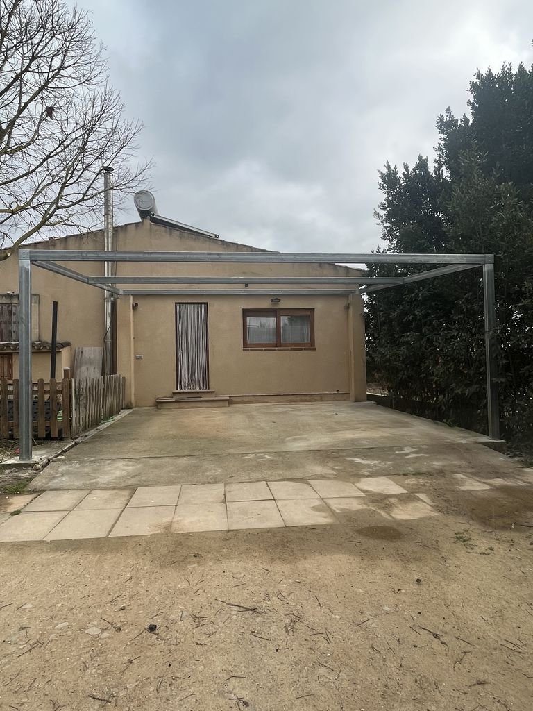Cochetera en construcción frente a una casa sencilla con ventanas y puerta, árboles a los lados, cielo nublado.