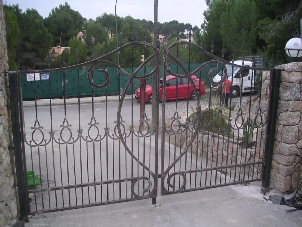 Valla de hierro con un corazón decorativo en el centro, en una entrada de un camino con plantas y vehículos estacionados al fondo.