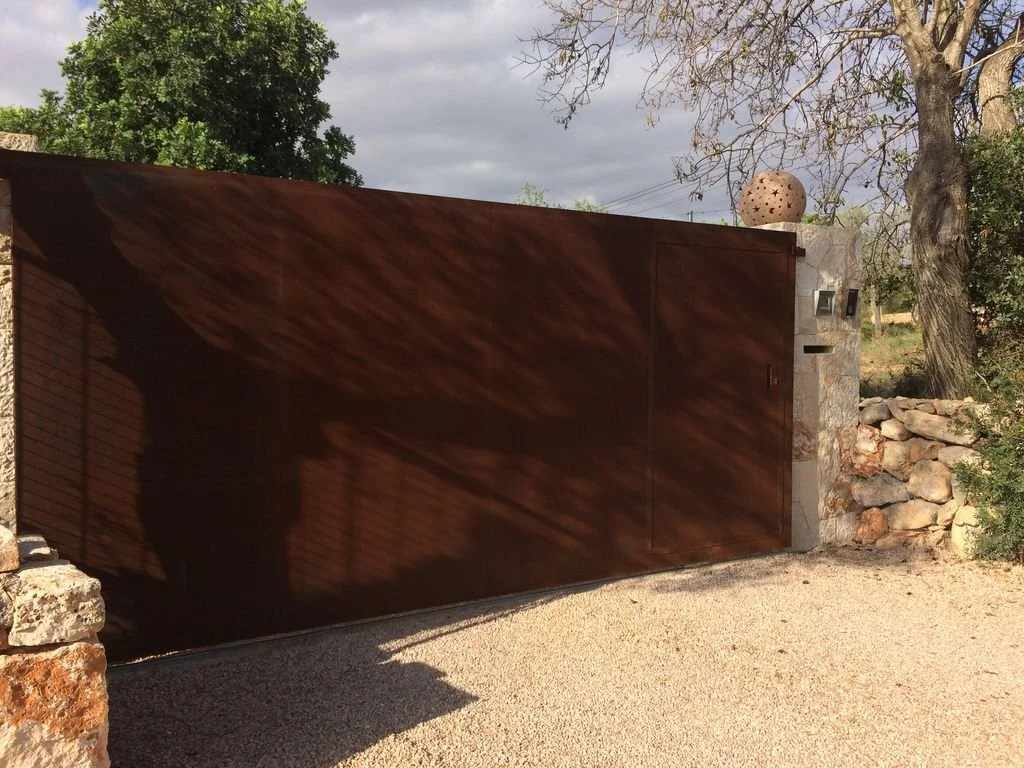 Puerta metálica grande de color marrón, con sombra proyectada de las ramas de un árbol sobre ella, en un entorno exterior con piedras y árboles.