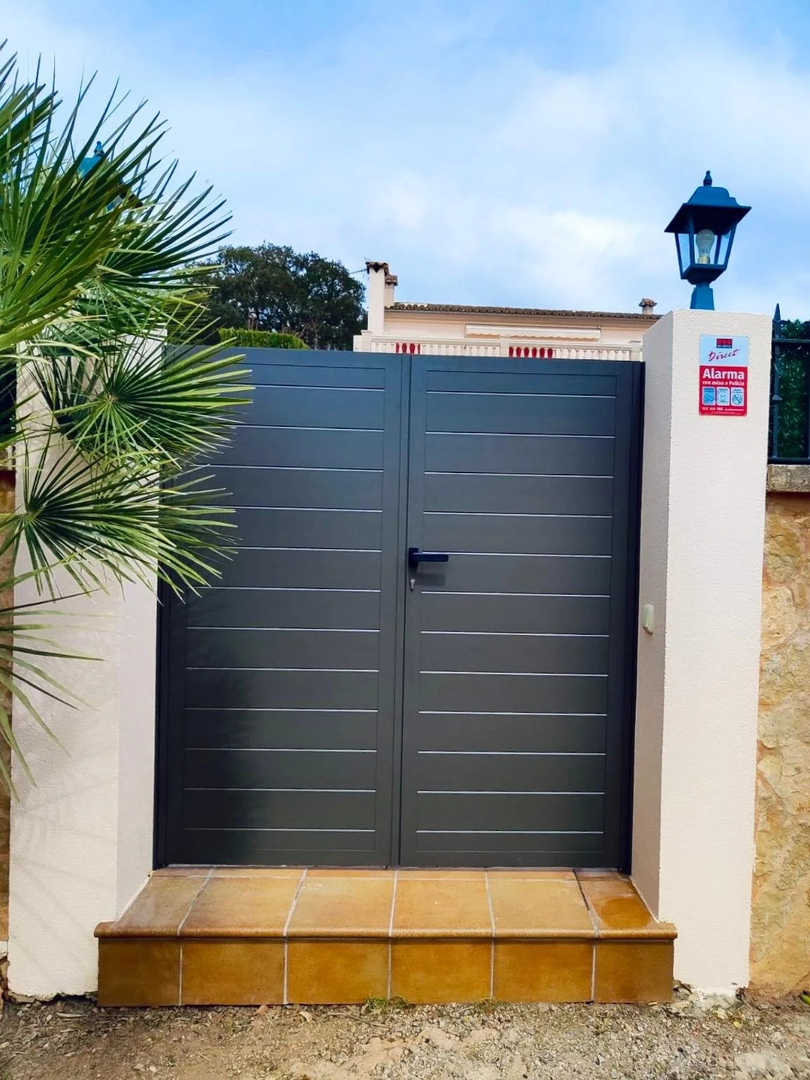 Puerta gris con cerradura en una entrada de vivienda, adornada con plantas verdes y una farola azul en la esquina derecha, y señal de alarma en la pared blanca lateral.