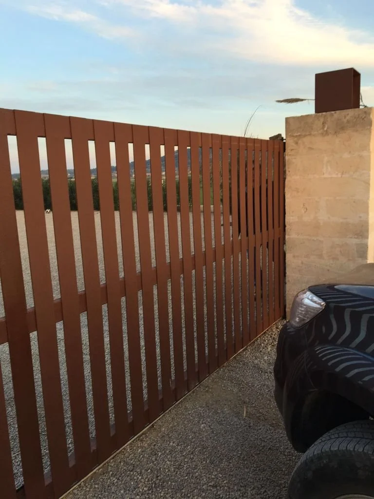 Una cerca de madera de color marrón, a la derecha hay una pared de piedra y parte de un automóvil negro.