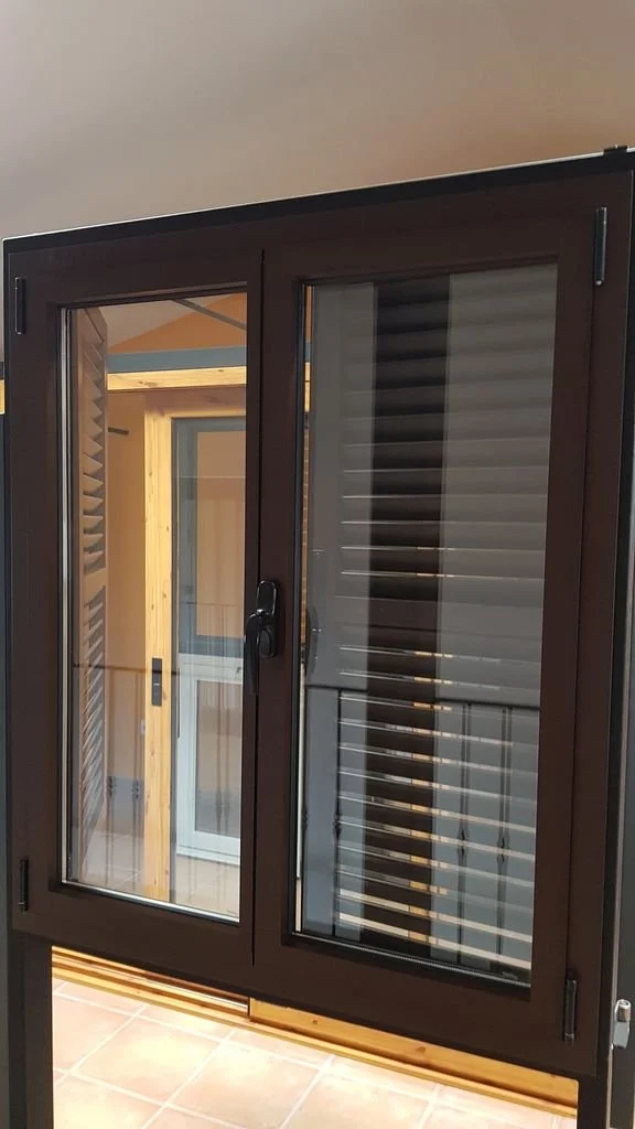Ventana de aluminio con marco oscuro y persiana en interior, en un espacio cerrado con suelo de baldosas.