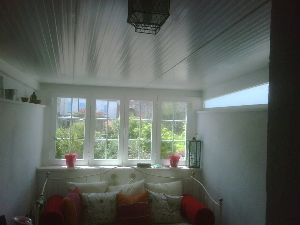 Habitación con ventanas grandes, sillón con cojines, plantas en la ventana y lámpara en la pared.