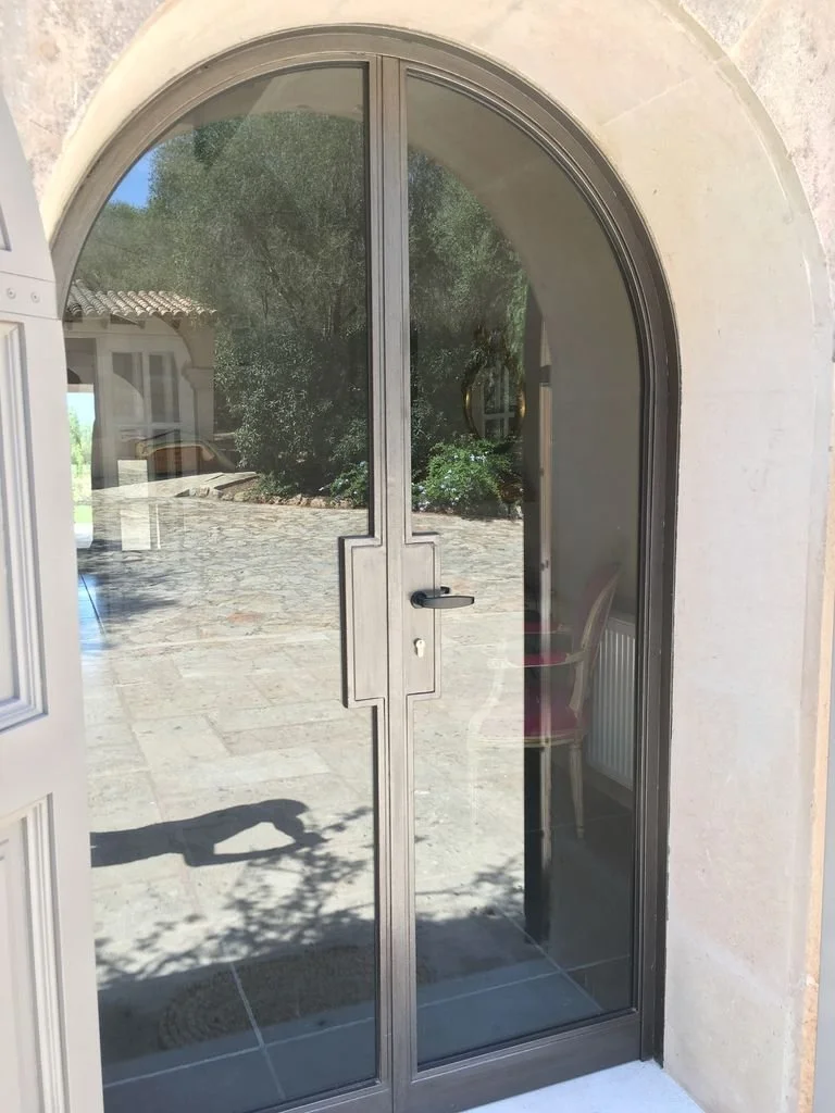 Puerta de vidrio doble con arco, situada en la entrada de un edificio, con vista al exterior y reflejo del jardín y las plantas.