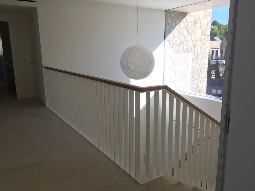 Vista del pasillo con barandal de madera y barandal de madera, lámpara de papel y ventana grande que muestra un edificio con piedra y árboles.