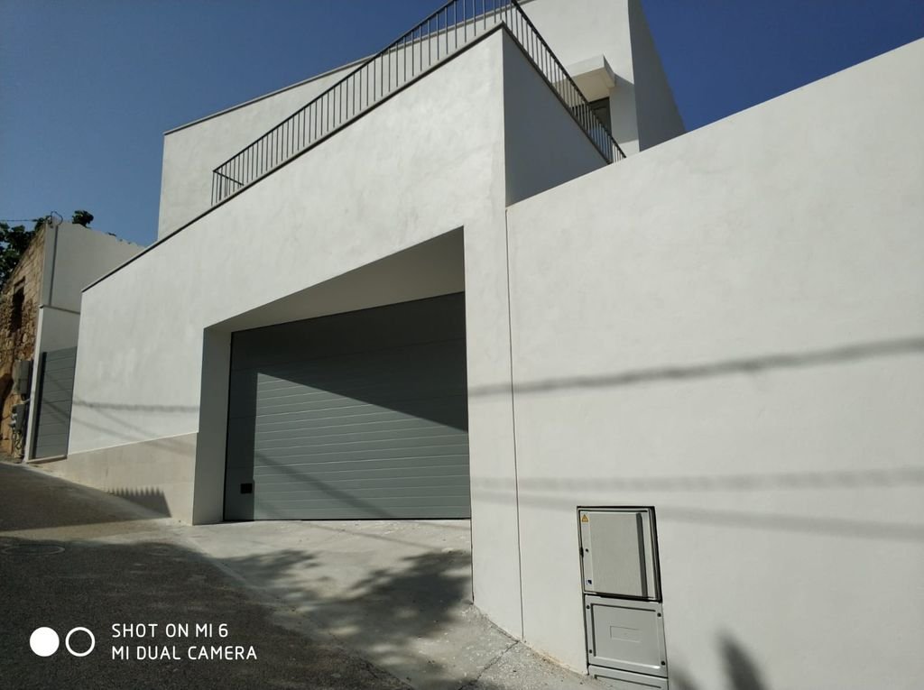 Edificio moderno con paredes blancas y una puerta de garaje gris, en una calle con sombra y cielo despejado.