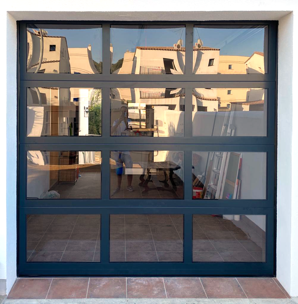 Puerta de garaje de vidrio con marco negro, reflejando las casas cercanas y el cielo, en un costado de una pared blanca.