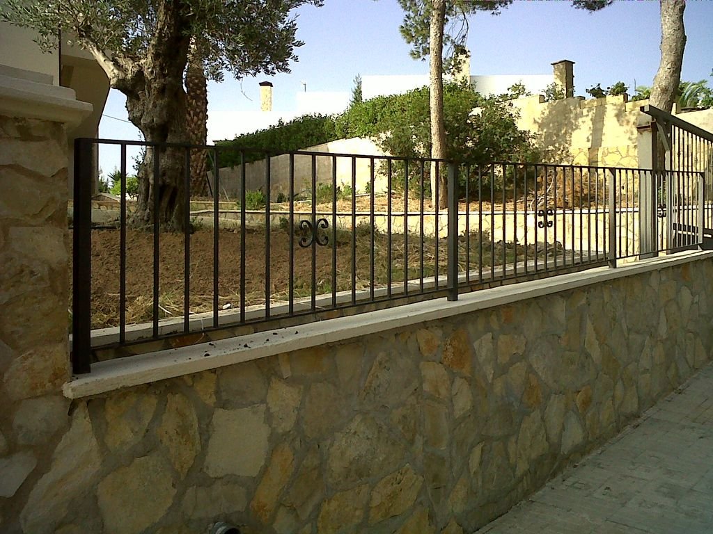 Barandal negro de metal sobre un muro de piedra, al fondo árboles y plantas en un área elevada.
