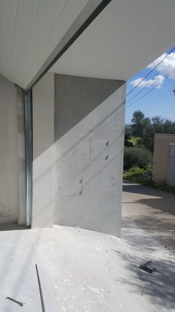 Vista exterior de una construcción en progreso, con paredes de hormigón y un suelo de cemento, bajo un cielo despejado y cables eléctricos en el fondo.