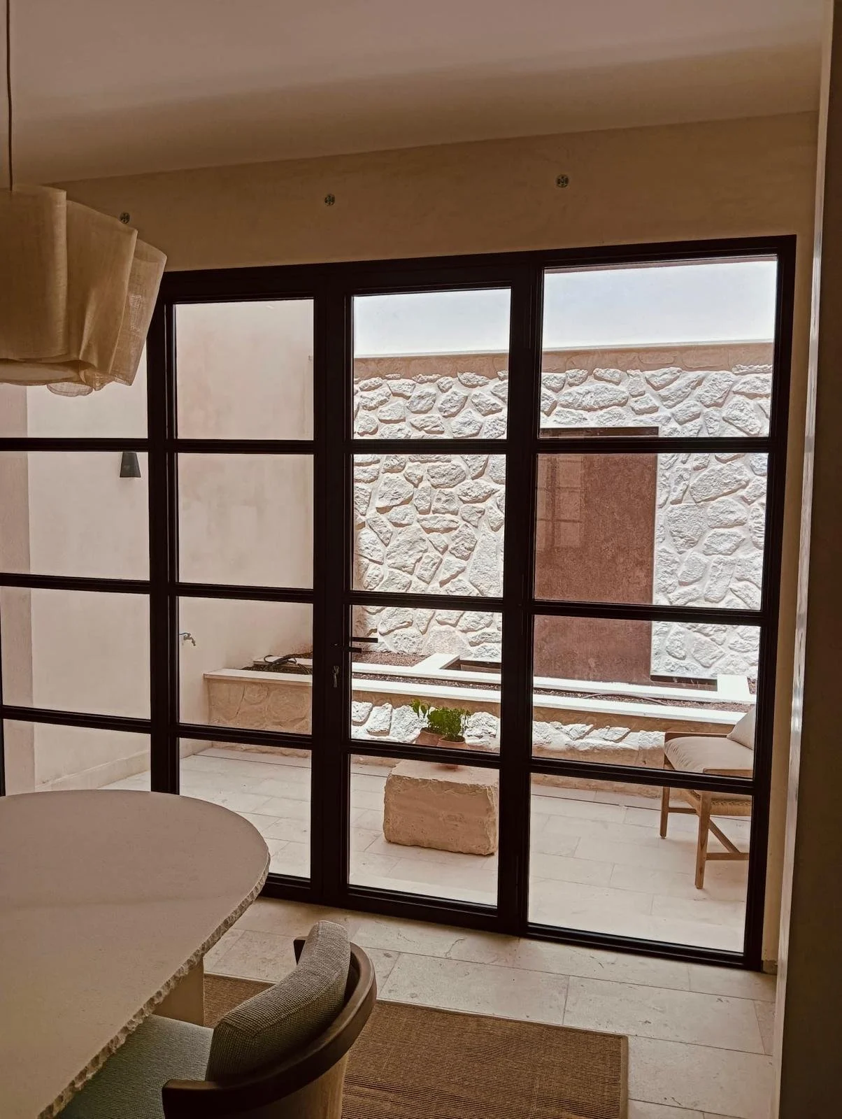 Vista de un patio interior con muros de piedra, una mesa y sillas, visto a través de una puerta de metal con vidrio.