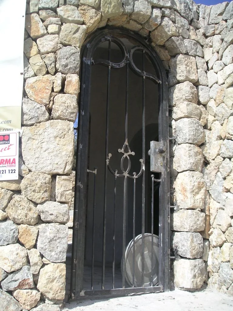 Puerta de hierro negro con rejas, enmarcada por piedras de muro, con candado y manija, en un muro de piedra.
