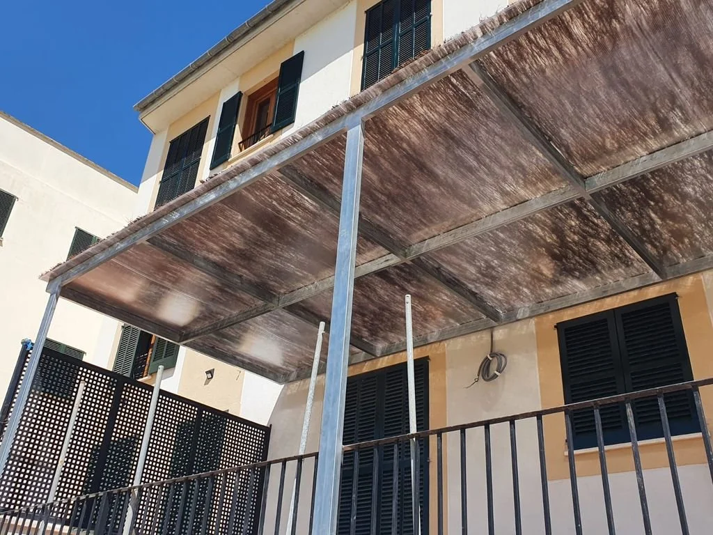 Perspectiva desde abajo de una estructura de alero de madera y metal en un balcón de un edificio, con ventanas con persianas negras.