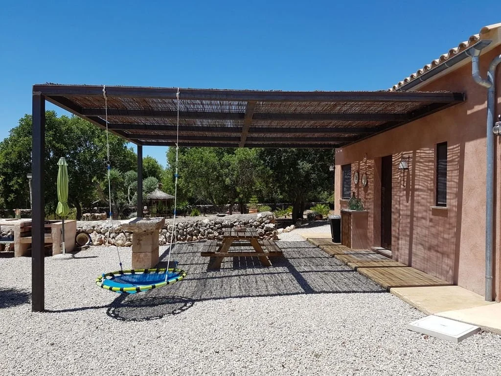 Porche cubierto con columpios y mobiliario de exterior en un patio con árboles y suelo de grava.