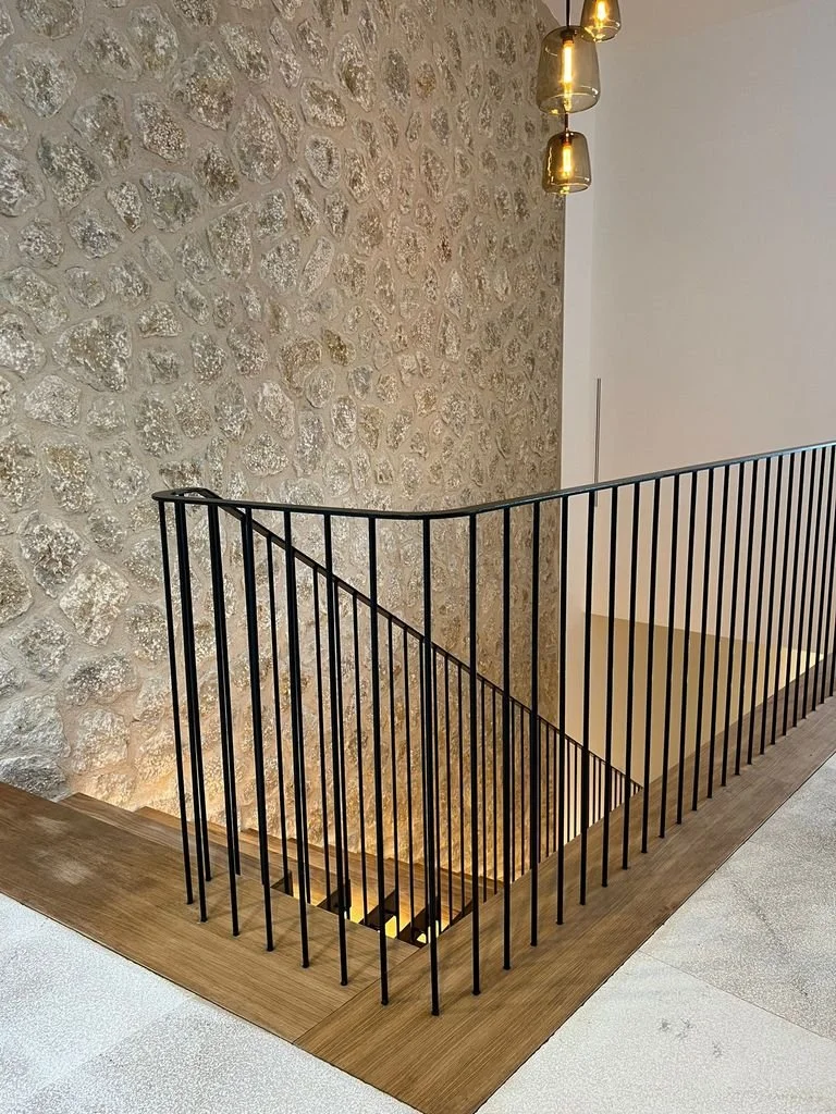 Escalera de madera con barandal de barras negras, pared de piedra en el fondo y lámparas colgantes doradas.