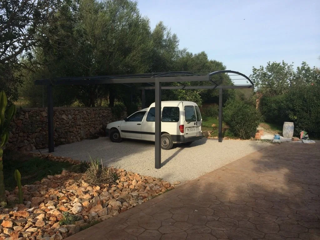 Una cochera techada con estructura de metal y una camioneta blanca estacionada en un área de grava, en un entorno rodeado de árboles y plantas.