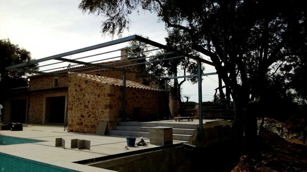Vista parcial de una casa en construcción con estructura metálica en un patio con piscina y adyacente un árbol grande en un día soleado.