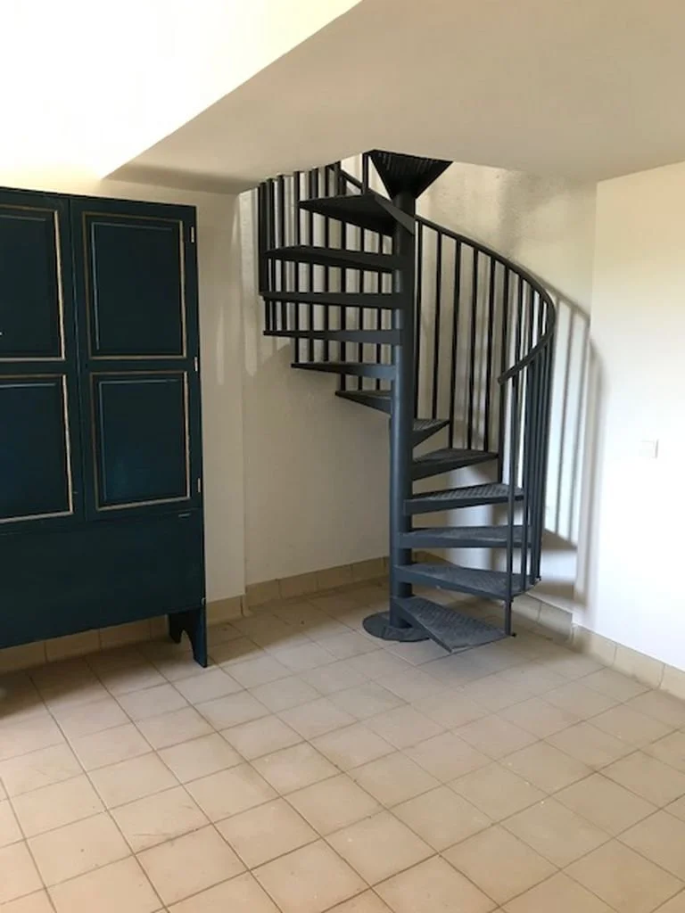 Escalera de caracol negra en una habitación con suelo de baldosas beige y un mueble azul a la izquierda.