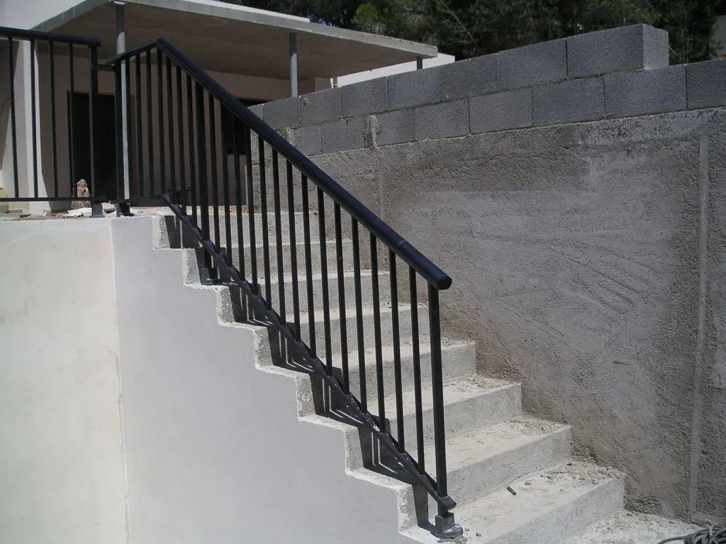 Escalera de concreto con barandal negro, en construcción o reparación en un exterior, con muro de bloques de cemento a un lado y estructura de techado en el fondo.