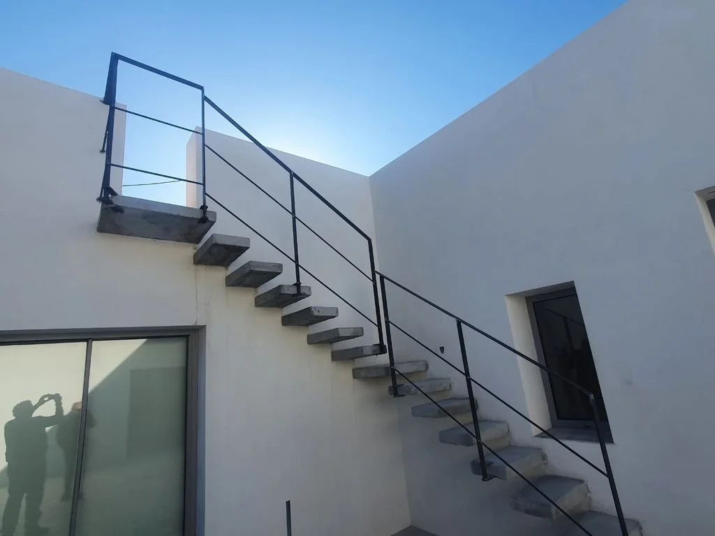 Escalera exterior de concreto con barandal negro en una vivienda moderna con paredes blancas y ventanas grandes, bajo un cielo despejado.