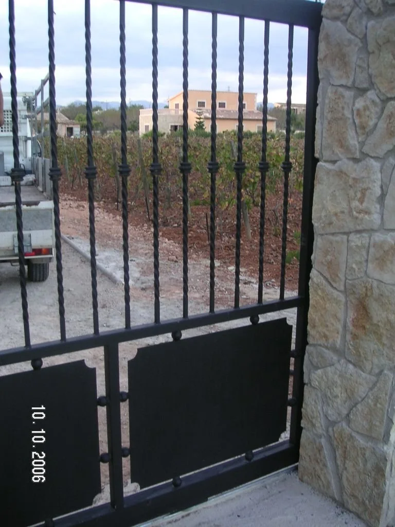 Barandal de metal negro frente a un terreno agrícola con plantas y casas al fondo, junto a una estructura de piedra.