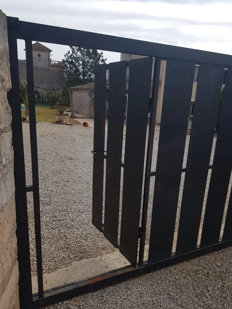 Puerta de rejas negra parcialmente abierta en un entorno exterior con suelo de grava y estructuras antiguas en el fondo