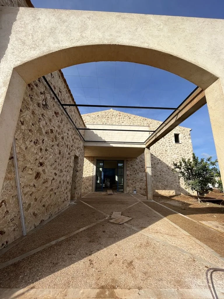 Construcción en progreso de una estructura de piedra y concreto, con arcos y paredes de piedra, en un ambiente exterior con cielo azul y sombra en el suelo.