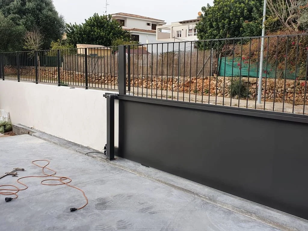 Una cerca metálica negra instalada en una barda blanca, con una puerta corrediza. En el fondo hay árboles y casas.