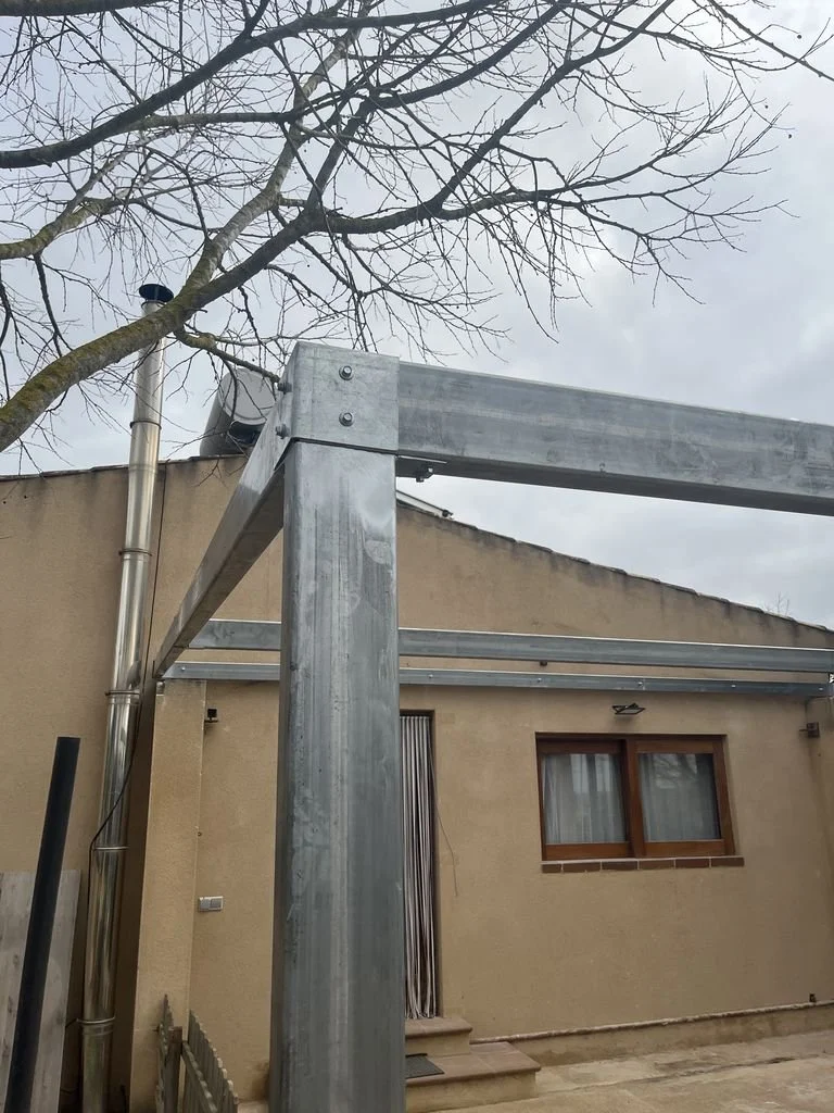 Estructura metálica en construcción frente a una casa con ventanas de madera y puertas, con árboles sin hojas en un día nublado.