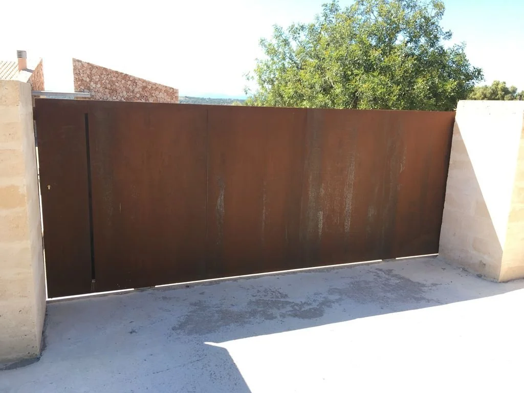 Puerta de metal oxidado en un muro de concreto, con árboles y un cielo despejado al fondo.