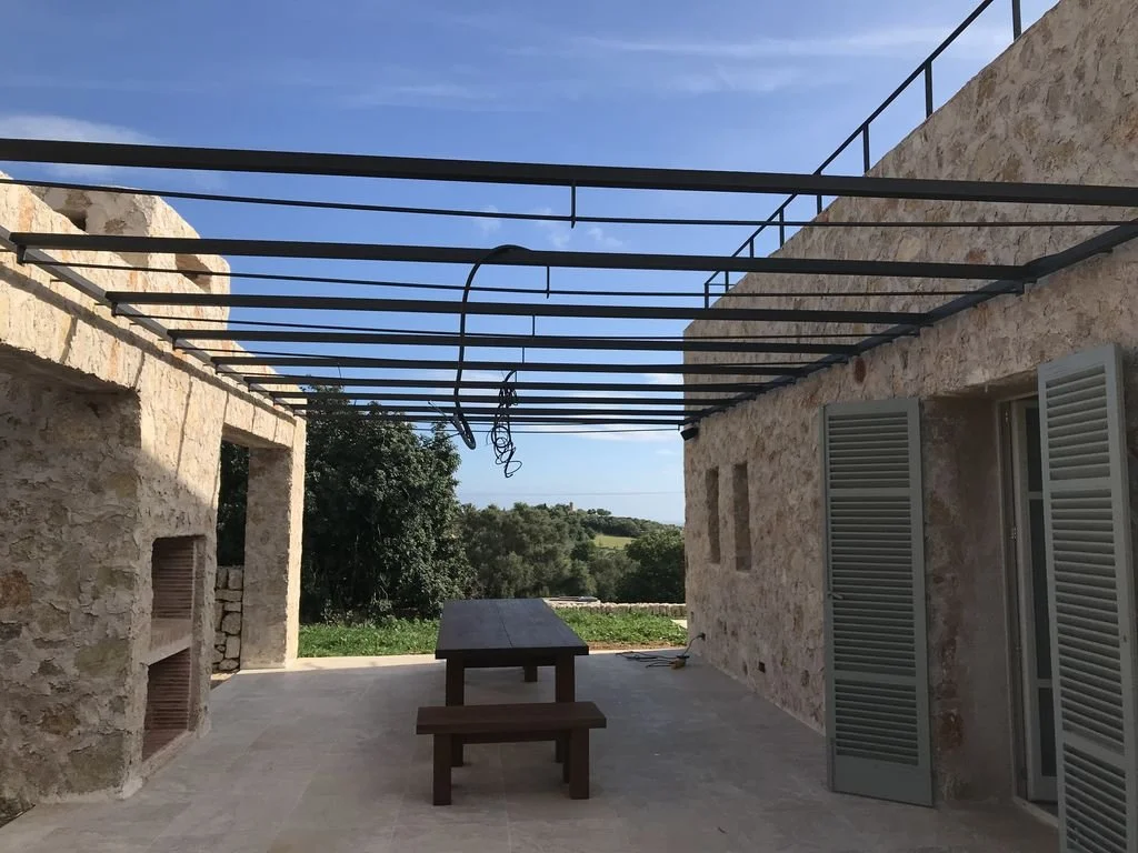 Terraza exterior de una casa en construcción con estructura de hierro para techo, paredes de piedra, ventanas con persianas de madera, mesa y banco de madera, y un fondo con paisaje natural y cielo despejado.