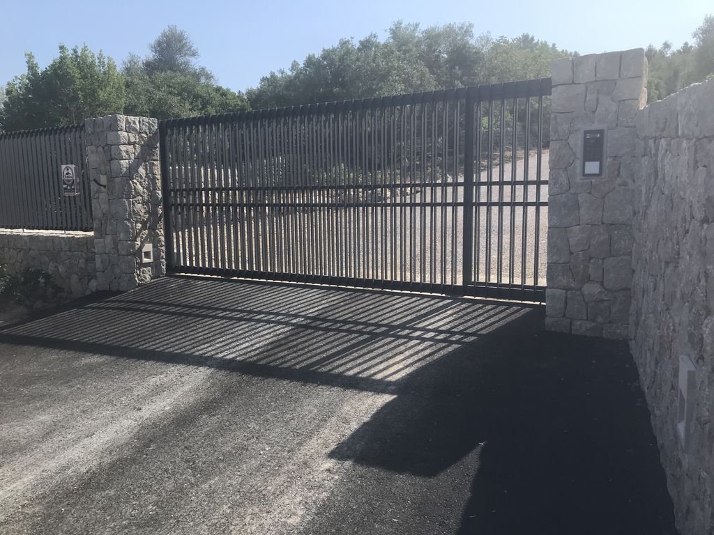 Portón de metal negro que se cierra con un muro de piedra a ambos lados, en un camino pavimentado, con sombra proyectada en el suelo.