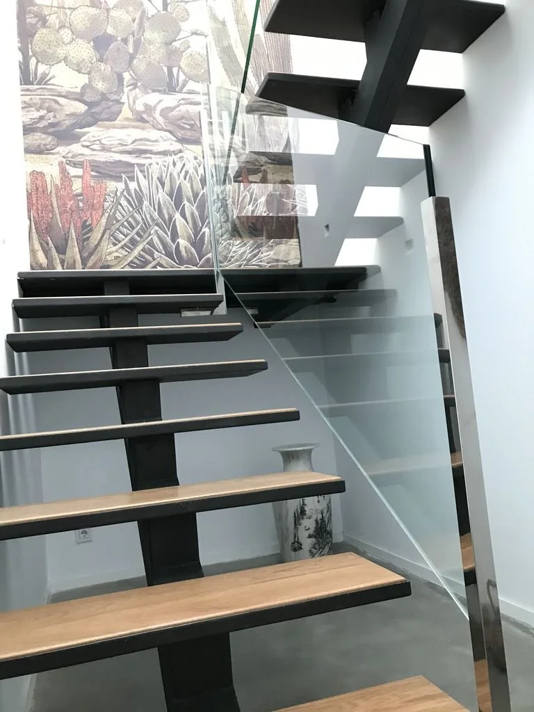 Escalera moderna con peldaños de madera y barandilla de vidrio y metal, en interior con mural de plantas y cactus.