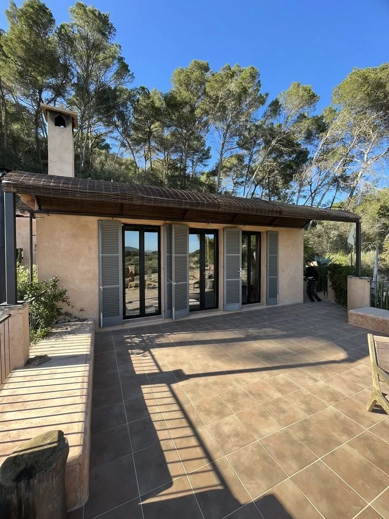 Terraza exterior con puertas corredizas de vidrio, ventanas con persianas, sombra de barandilla en el piso, rodeada de árboles y cielo azul.