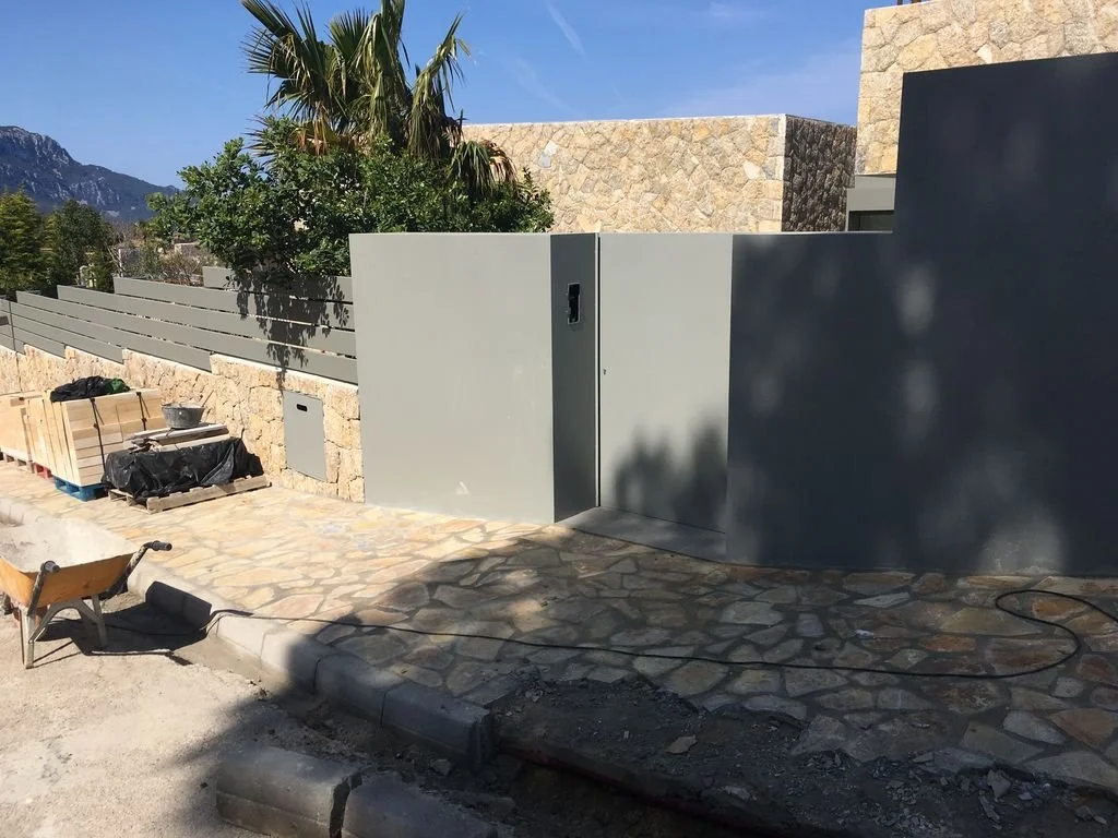 Pared de bloques de piedra con puerta moderna y muro gris, rodeado de vegetación y construcción en progreso con herramientas y materiales.