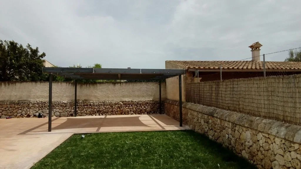 Patio con baldosa, estructura de techo para coche, césped y muro de piedra con valla de malla.