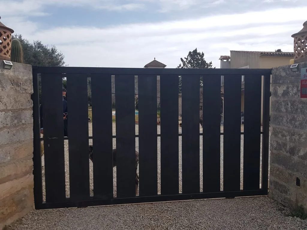 Puerta de metal negra con listones verticales, entre muros de piedra, en un acceso de una propiedad.