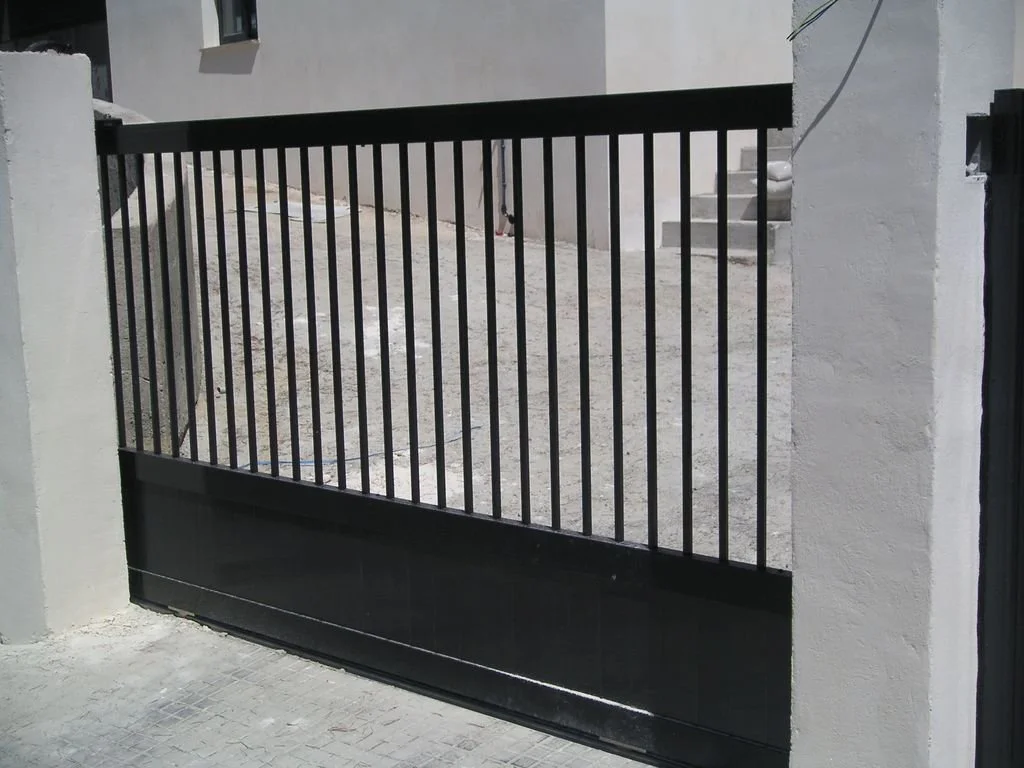 Puerta metálica negra con rejas en una entrada de casa, con escalera y muro de fondo.