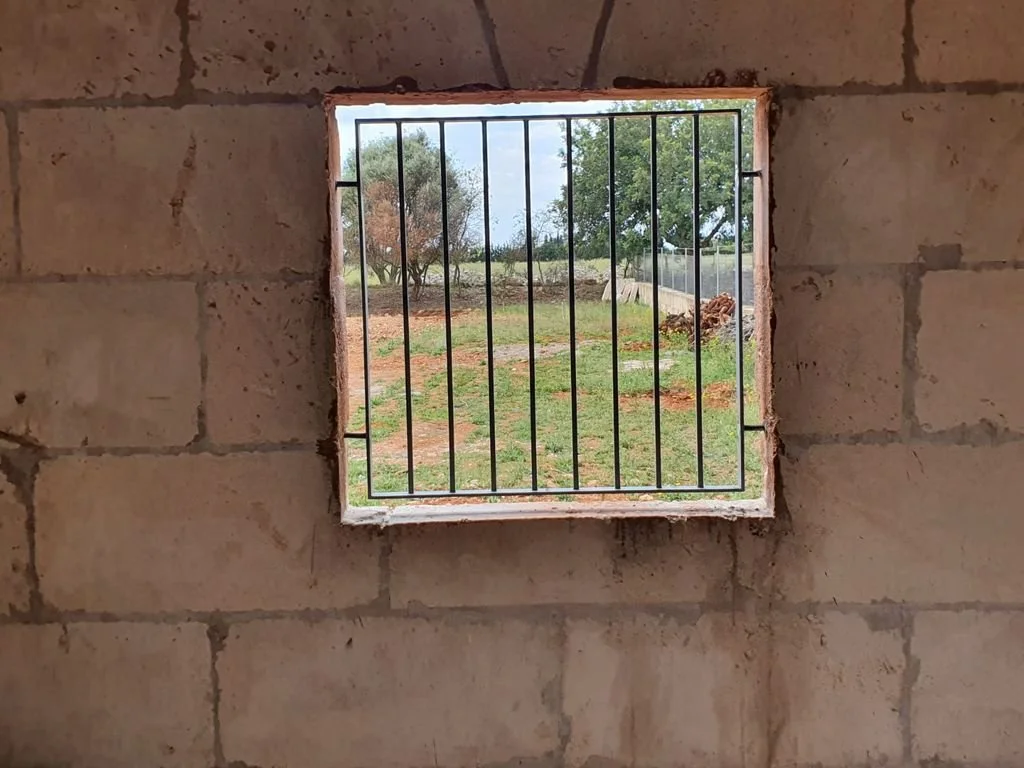 Ventana en una pared de bloque de concreto con barrotes de hierro, vista a un área de campo con árboles y césped.