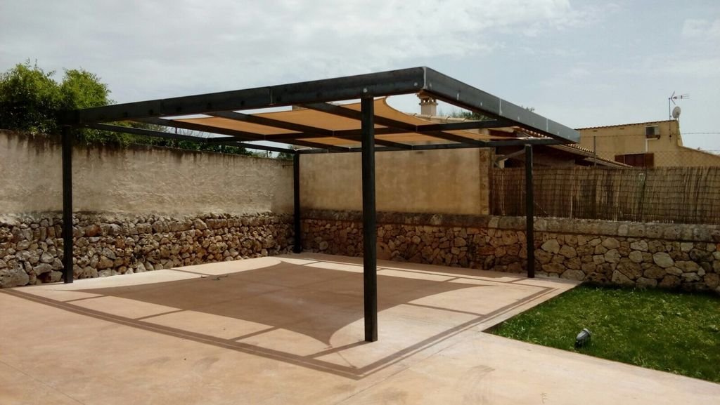 Estructura de techo con columnas en un patio, sombra proyectada y cercado de piedra en el fondo.