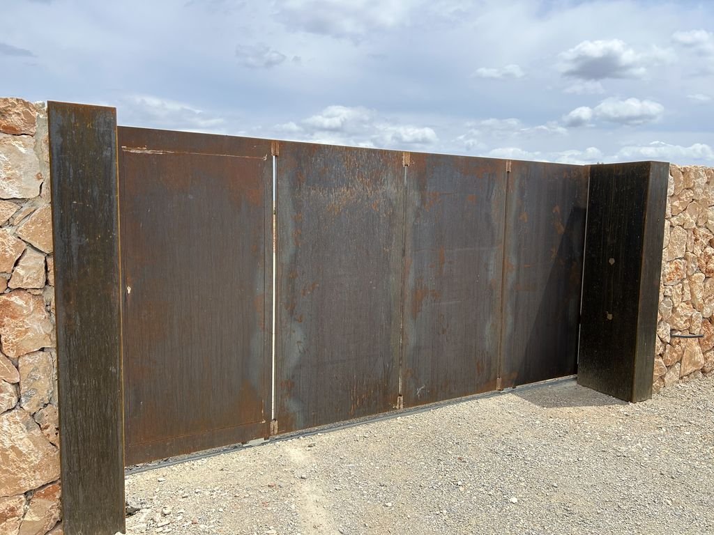 Una puerta grande de metal oxidado con dos postes de madera a los lados, ubicada frente a una pared de piedra y un cielo nublado.