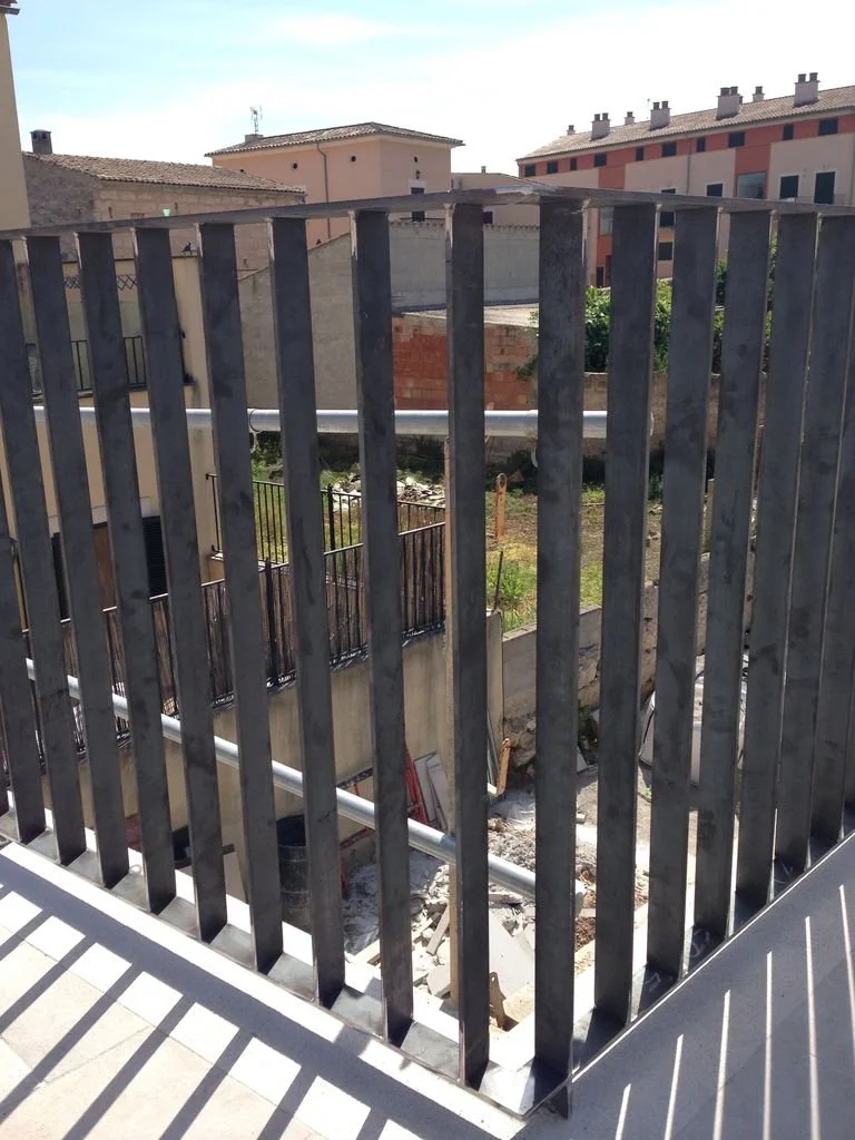 Vista de una construcción en proceso con barras de hierro y bloques de cemento en un balcón, con edificios residenciales en el fondo.