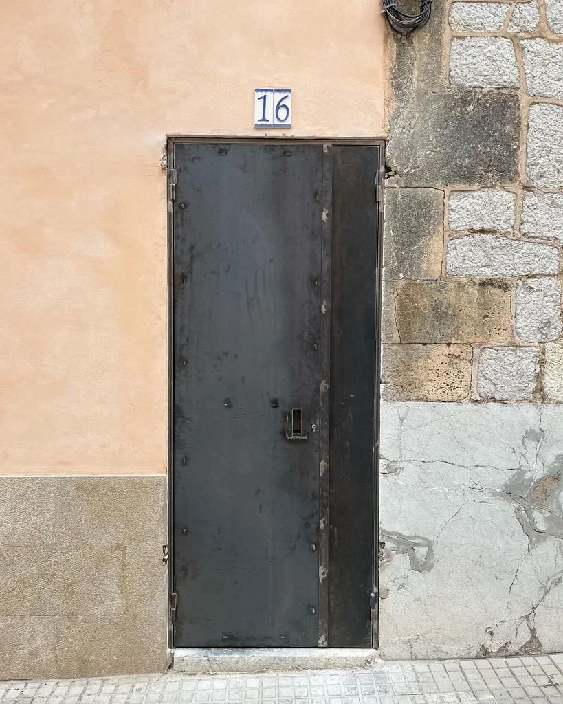 Puerta de metal negra con número 16 en azulejo azul y blanco en la pared de un edificio de piedra y estuco.