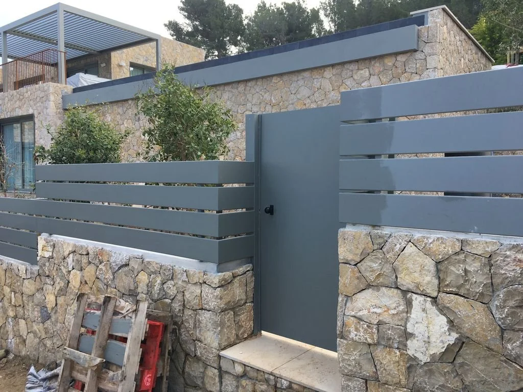 Muro de piedra con puerta y reja de madera pintada de gris, en una casa moderna con terraza y vegetación.