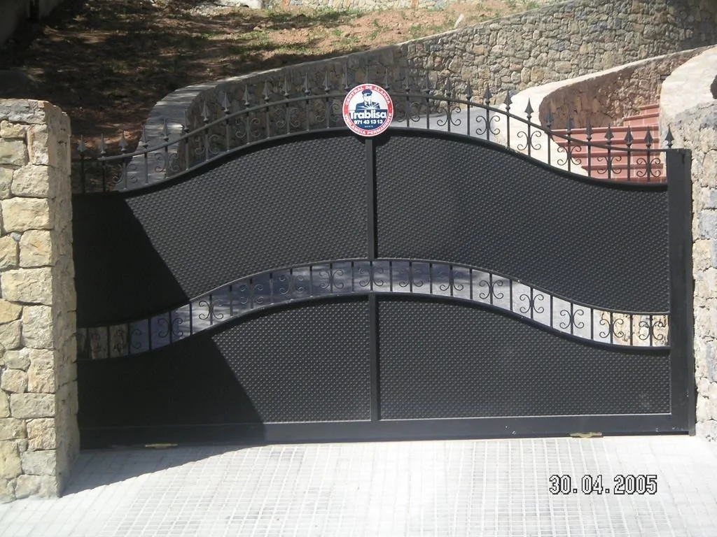 Puerta de metal negra con diseño en curvas y detalles en hierro, rodeada de muros de piedra.