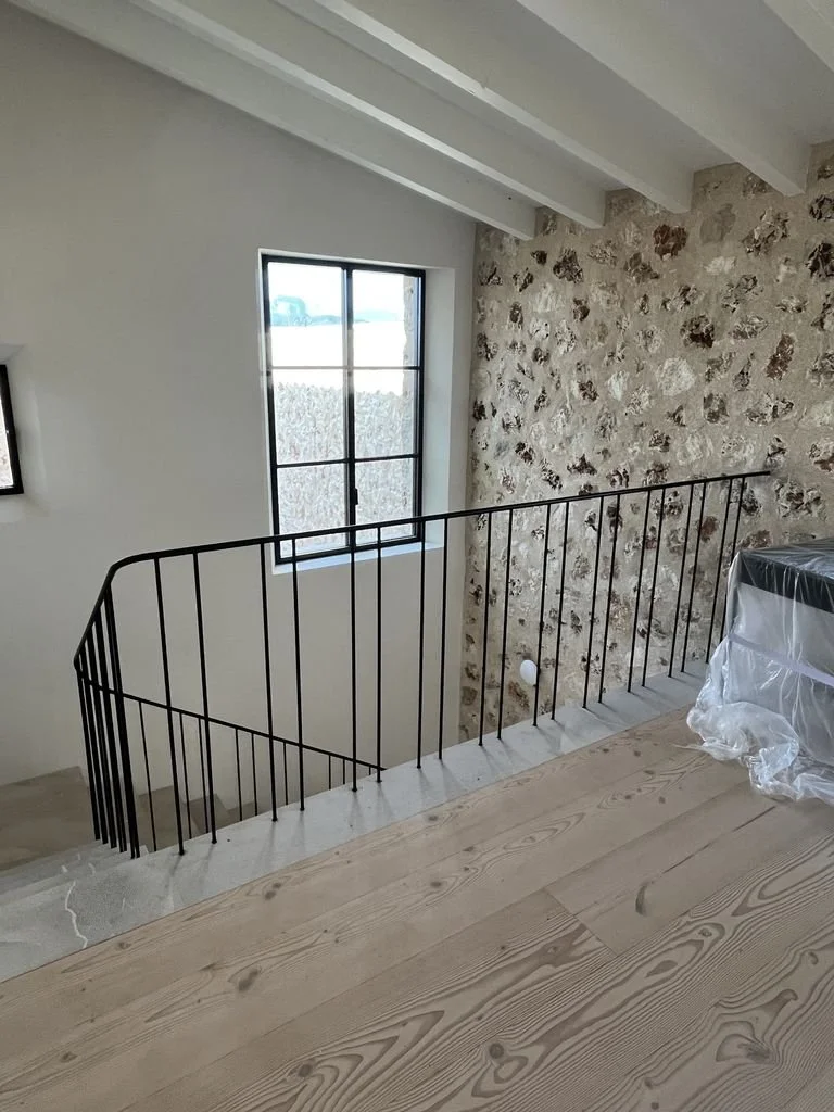 Interior de una casa en construcción con una barandilla negra de metal, ventanas de vidrio y muros de piedra rústica, vista desde el segundo nivel hacia las escaleras.